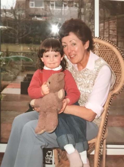 Linda Owst sitting on a chair in front of a window with her granddaughter sitting on her knee holding a teddy bear.